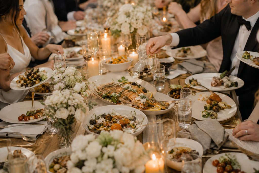 Family-style wedding table with shared platters, plated portions, candles, floral centerpieces, and guests dining together