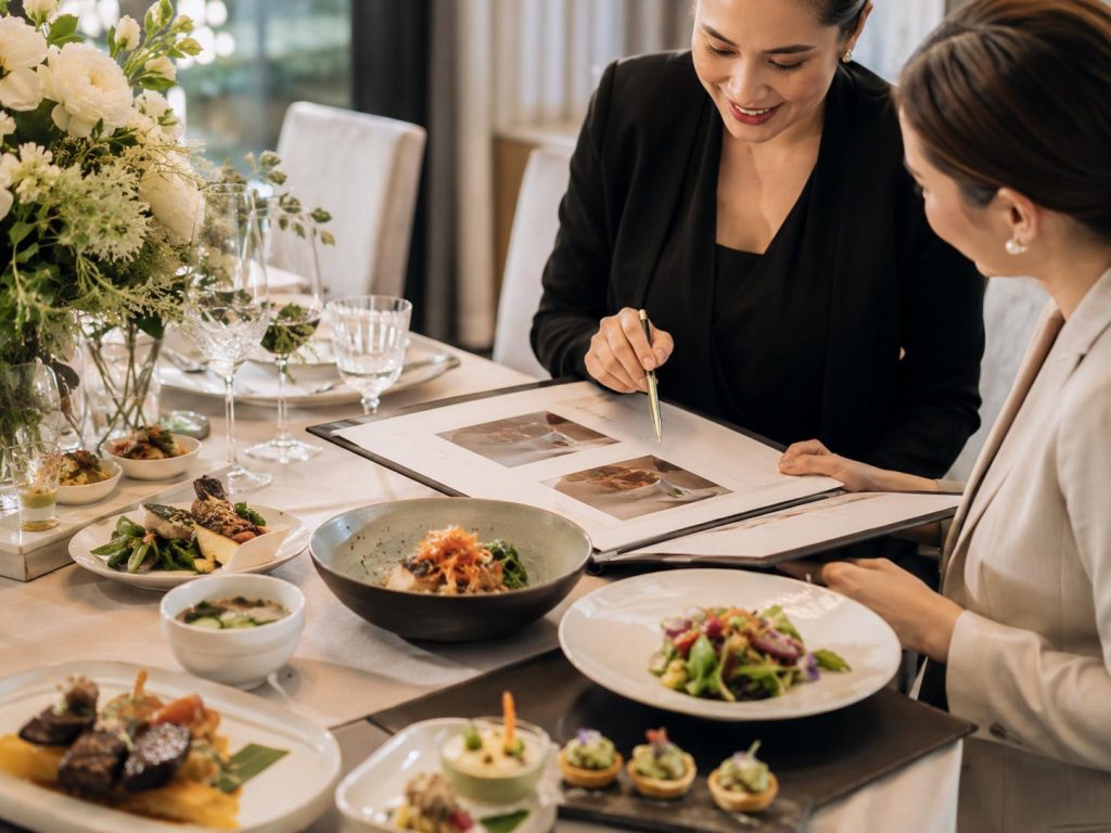 Catering specialist reviewing menu options with a client beside plated tasting dishes, glassware, floral accents, and menu samples