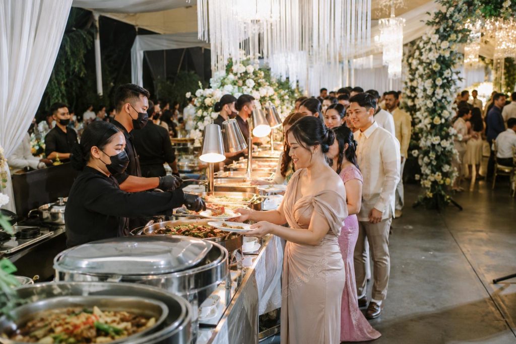 Guests line up at an elegant buffet station while catering staff serve dishes at a large wedding reception