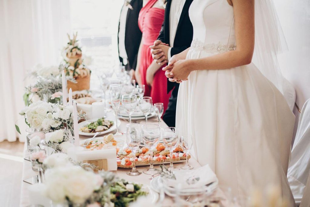 Bride and guests stand beside a styled event table with plated dishes, glassware, and floral accents